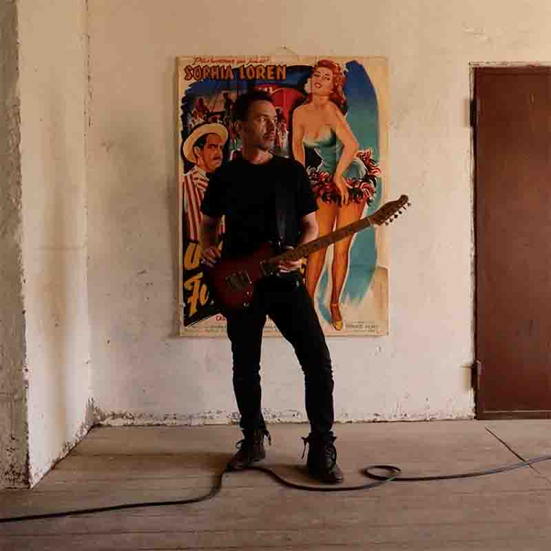 Ibiza Camping Site on the beach Multiinstrumentalist and Music Producer Likewolf playing an electric guitar with a sunburst finish. Likewolf is standing in a room with a white wall that has a colorful poster featuring “Sophia Loren”. He is dressed in a black t-shirt, black pants, and black shoes. A black cable runs from the guitar to an amplifier, set on a wooden floor.
