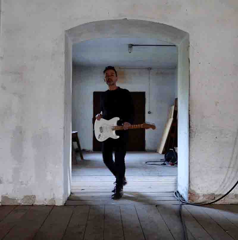 Ibiza Camping Site on the beach Multiinstrumentalist and Music Producer Likewolf standing in an archway, holding a white electric guitar. Likewolf is dressed in a black long sleeve shirt and black pants. A wooden floor underfoot has a cable running across it, hinting at the musical setting.